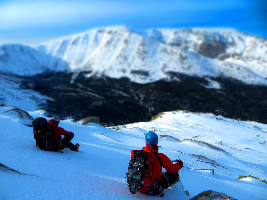 A climber high on a snow-covered mountain ridge under a wide alpine sky.